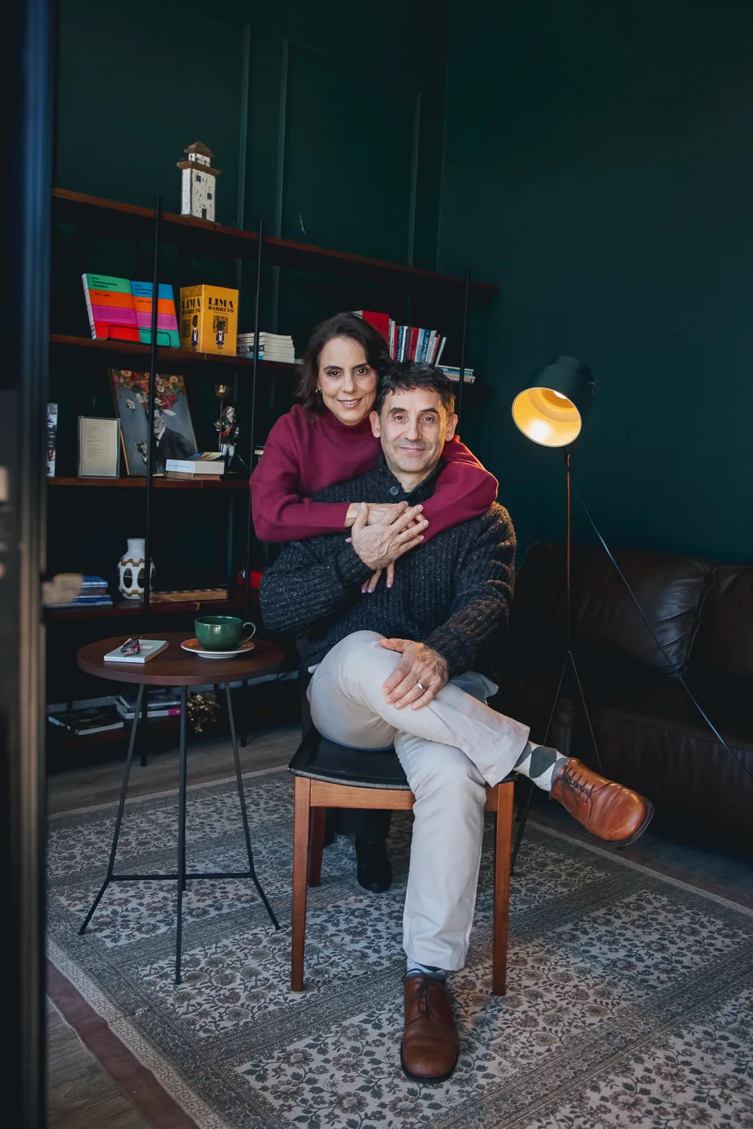 Sandra Caselato abraçando Yuri Haasz, sentado, numa sala com estante de livros e luminária